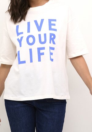 Henkilö, joka pukeutunut valkoiseen, lyhythihaiseen paitaan, jossa on rohkea sininen teksti "LIVE YOUR LIFE", yhdistetty tummansinisiin farkkuihin, yksinkertaisen taustan edessä.