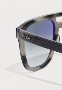 Ochelari Ray-Ban cu ramă gri degradé, lentile cu nuanță albastră și un logo proeminent pe braț. Textură netedă, formă pătrată modernă.