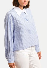 Chemise à rayures bleues avec un col blanc, présentant de légères lignes verticales et une coupe ample. Confectionnée en tissu léger.