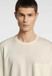 Lys beige oversized t-skjorte laget av mykt stoff, med rund hals og brystlomme på venstre side. Glatt tekstur.