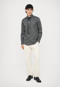 Czarna koszula w wzory z czarnym skórzanym krawatem, w połączeniu z kremowobiałymi spodniami i czarnymi eleganckimi butami. Model stoi na białym tle.