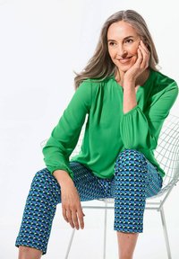 Blusa verde con maniche lunghe e polsini arricciati, abbinata a pantaloni capri blu e verde a fantasia con forme triangolari. Sfondo di una sedia bianca.