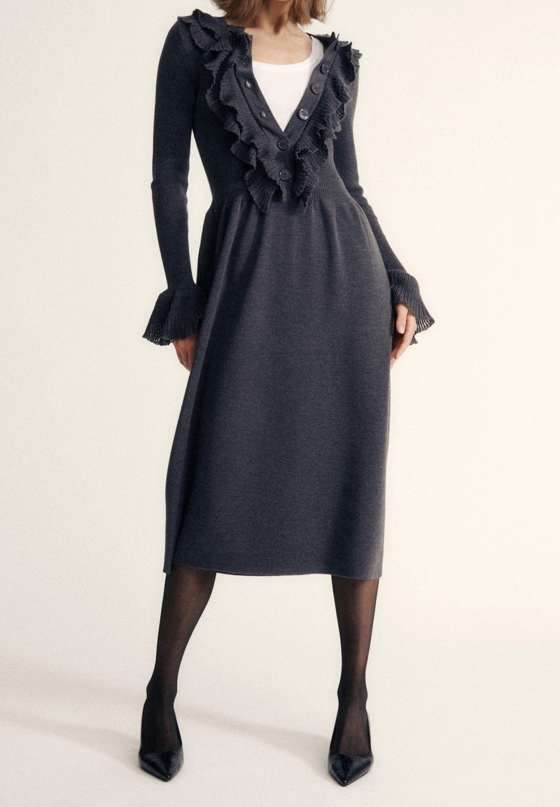 Femme portant une robe midi gris foncé avec un col en V à volants et des poignets, des collants noirs transparents et des escarpins noirs à bout pointu, debout devant un fond clair.