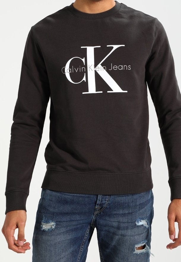 Homme portant un sweat-shirt noir Calvin Klein Jeans avec un grand logo blanc "cK" et un jean bleu délavé.