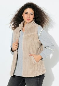 Gilet en peluche beige avec texture côtelée verticale, col montant, fermeture éclair et poches latérales. Superposé à une chemise à manches longues grise.