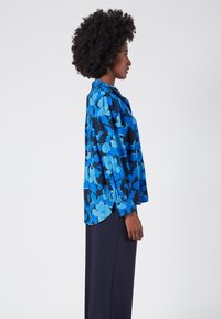 Niebieska kwiatowa bluzka z długim rękawem, wykonana z lekkiej tkaniny, z kołnierzykiem zapinanym na guziki i zaokrąglonym dołem, zestawiona z ciemnymi spodniami.