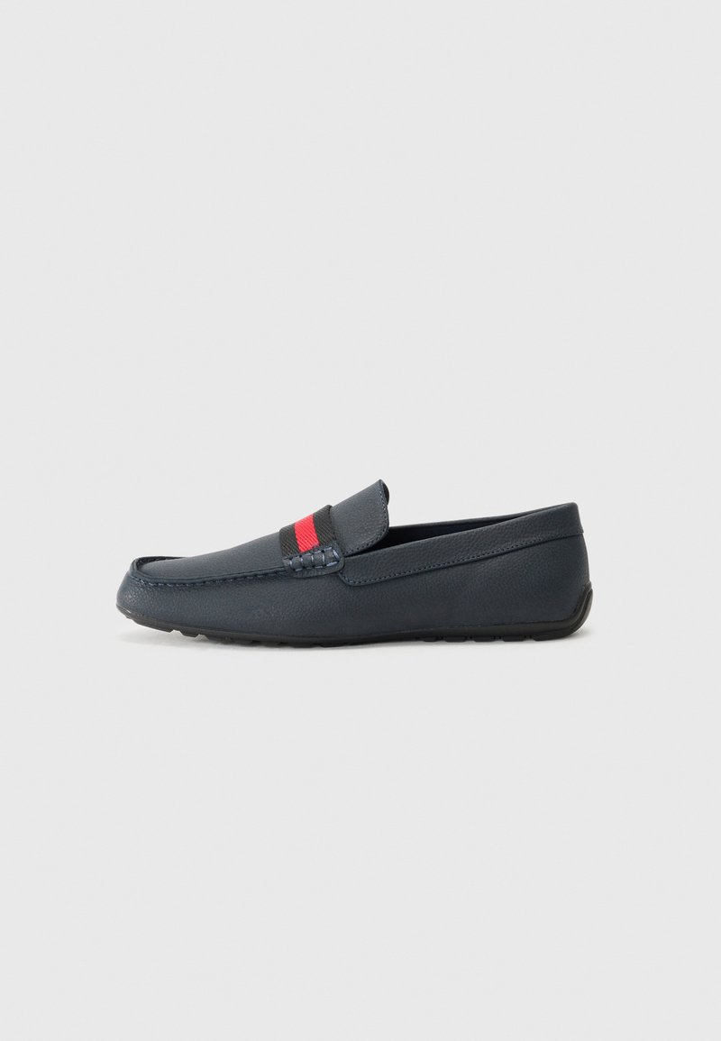 Sapatilha moccasin azul-marinho feita de pele texturizada, com uma faixa contrastante em vermelho e preto, ponta arredondada e sola de borracha plana.