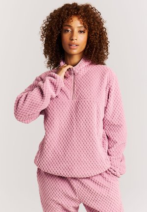 Vrouw met krullend haar, gekleed in een roze fleece trui met rits en bijpassende broek, staand tegen een effen lichte achtergrond.