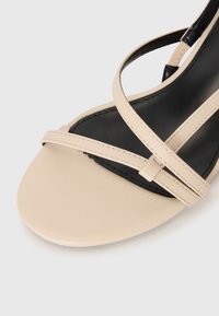 Beige lærsandale med tværsnørret design, sort indersål, glat tekstur og rund tåform. Elegant og minimalistisk stil.