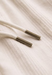 Gros plan de cordons en tissu blanc avec embouts métalliques reposant sur un tissu beige texturé.