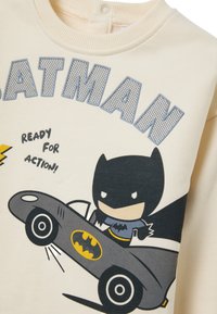 Hellecreme Sweatshirt mit einem Cartoon-Batman, der ein graues Auto fährt. Der Text lautet "BATMAN" und "BEREIT FÜR DEN EINSATZ!" in Blau und Schwarz.