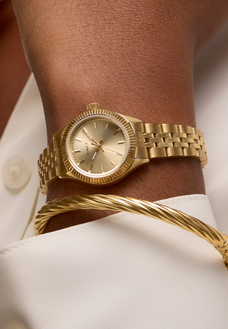 Montre ton or avec une lunette texturée et un bracelet à maillons métalliques, dotée d'un cadran or clair et de fines marques des heures, portée au poignet.