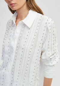 Witte knoopshirt met een gestructureerde stof met verticale strepen en verspreide zilveren versieringen voor een decoratief effect.