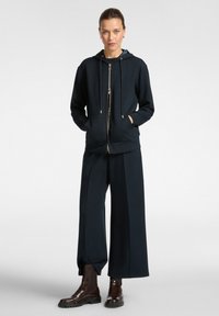 Felpa blu con cerniera e tasche, abbinata a pantaloni larghi blu. Presenta lacci e una texture morbida. Completato con stivaletti brown alla caviglia.