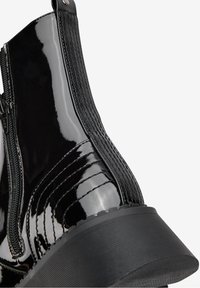 Acercamiento de una bota negra de charol con cremallera y tacón texturizado sobre un fondo blanco.