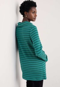 Zaļš svītrots garo piedurkņu džemperis ar kapuci, ar faktūrētu adījuma dizainu un sānu kabatām, uzvilkts virs tumšām biksēm.