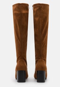 Ett par höga bruna mockastövlar med klackar sedd bakifrån mot en vit bakgrund.