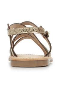 Vista trasera de sandalias de cuero marrón con correas metálicas decorativas, detalles texturizados y una suela plana.