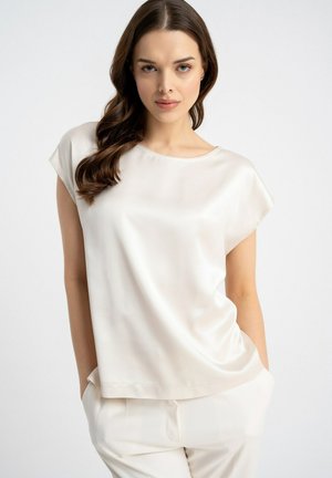 Młoda kobieta z falującymi brązowymi włosami, ubrana w białą satynową bluzkę z krótkim rękawem i dopasowane białe spodnie, stojąca z rękami w kieszeniach.