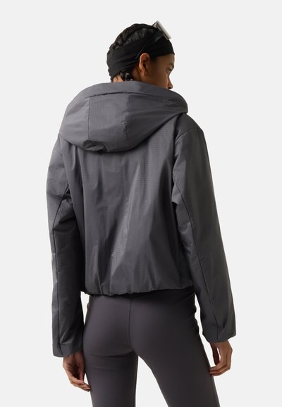 Osoba ubrana w ciemnoszarą kurtkę z kapturem i dopasowane ciemne legginsy, stojąca tyłem na tle jednolitego jasnego tła.