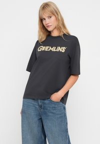 Pelēka kokvilnas t-kreklu ar īsām piedurknēm, kurā uz krūtīm liela bēša burtiem ir uzraksts "GREMLINS". Liela izmēra piegriezums.