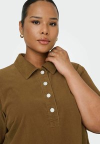 Chemise en velours côtelé marron avec col, ornée de boutons blancs et d'un tissu texturé. Une personne tient sa main près du col.