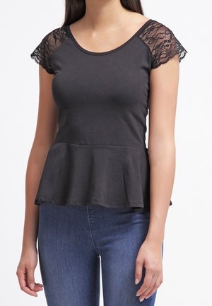 Vrouw die een zwart topje met korte mouwen, kanten schouders en een peplum zoom draagt, gecombineerd met een blauwe spijkerbroek, tegen een witte achtergrond.