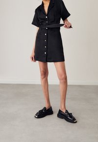 Robe noire à manches courtes avec fermeture à boutons et ceinture à la taille, confectionnée dans un tissu lisse. Portée avec des chaussures compensées noires ornées d'un nœud.
