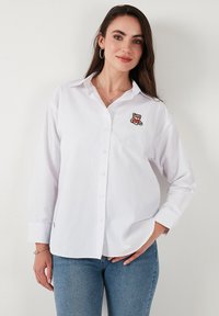 White long-sleeve button-up shirt, with an embroidered bear design on the left chest. Soft fabric, tailored collar, and straight cut.