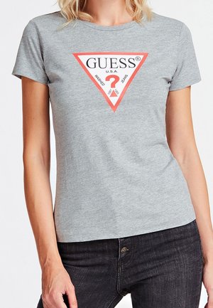 Femme portant un t-shirt gris Guess avec un logo triangulaire rouge et blanc et un jean noir, debout devant un fond uni.