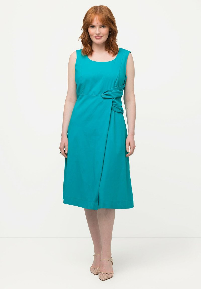 Donna dai capelli rossi che indossa un vestito senza maniche di colore teal, lungo fino al ginocchio, con dettagli drappeggiati sui lati, e tacchi a punta beige, in piedi su uno sfondo bianco.