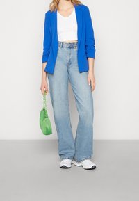 Kvinna iklädd en blå blazer, vit crop top, ljusblå vida jeans, vita sneakers, hållande en liten grön handväska med kedja.