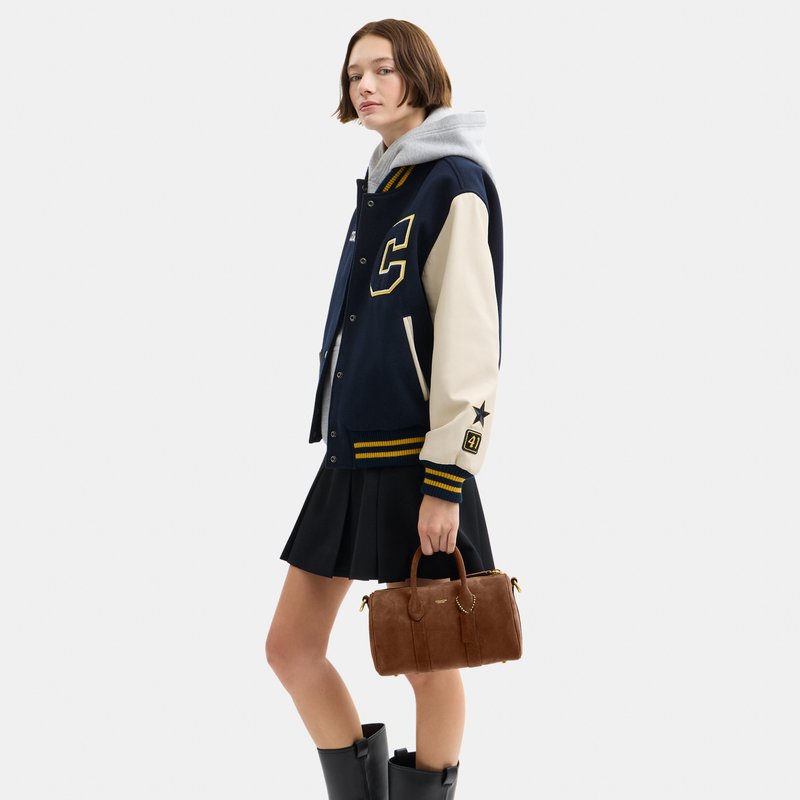 Jeune femme portant une veste varsity bleu marine et une jupe noire, tenant un sac à main marron, debout et regardant devant elle.