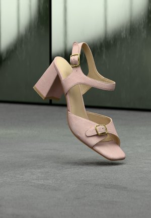 Pinkfarbene Wildleder-Sandalette mit hohen Absätzen, zwei verstellbaren goldfarbenen Schnallenriemen, Blockabsatz und offenem Zehenbereich auf grauer Oberfläche vor unscharfem Hintergrund.