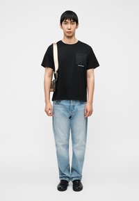 Μαύρο T-shirt με κοντά μανίκια, διακοσμημένη τσέπη και λογότυπο, σε συνδυασμό με ανοιχτό μπλε τζιν παντελόνι με ίσια γραμμή και μαύρα αθλητικά παπούτσια.