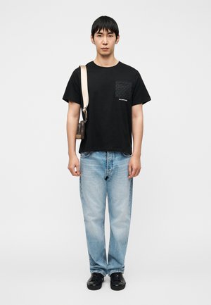 Μαύρο T-shirt με κοντά μανίκια, διακοσμημένη τσέπη και λογότυπο, σε συνδυασμό με ανοιχτό μπλε τζιν παντελόνι με ίσια γραμμή και μαύρα αθλητικά παπούτσια.