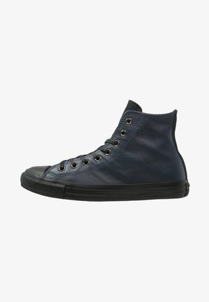 Baskets montantes en cuir noir avec semelle en caoutchouc noire, embout noir et lacets noirs passés à travers huit œillets métalliques sur fond blanc.