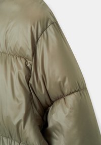 Gros plan sur la manche d'une veste matelassée vert olive montrant un tissu lisse et brillant avec des coutures horizontales.