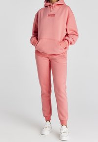 Persona vistiendo un conjunto a juego de sudadera con capucha rosa y joggers con bolsillos frontales y zapatillas blancas, de pie contra un fondo liso.