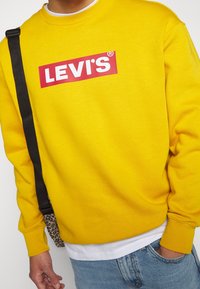 Gelbes Sweatshirt mit einem roten Levi's-Logo auf der Vorderseite. Mit gerippten Bündchen und einem klassischen Rundhalsausschnitt, präsentiert mit einer gemusterten Tasche.