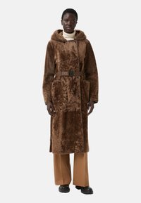 Hnědý umělý kožešinový kabát s kapucí, který má páskový pas, dvě přední kapsy a boční rozparky, v kombinaci s béžovými kalhotami a černými botami.