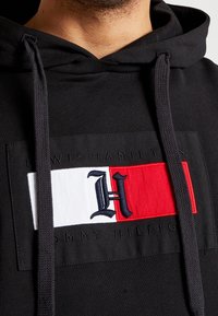 Zwarte hoodie met een rechthoekige patch. De patch heeft een rood-wit-rood ontwerp met een geborduurde "H" en de tekst "TOMMY HILFIGER".