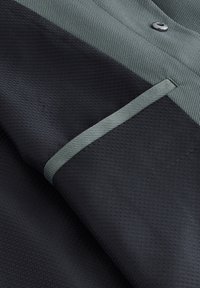 Primer plano de tela texturizada negra y gris con un botón y una pequeña costura horizontal en el bolsillo.