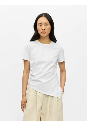 Femme aux longs cheveux foncés portant un haut asymétrique blanc à manches courtes et un pantalon beige à jambes larges, debout devant un fond blanc uni.