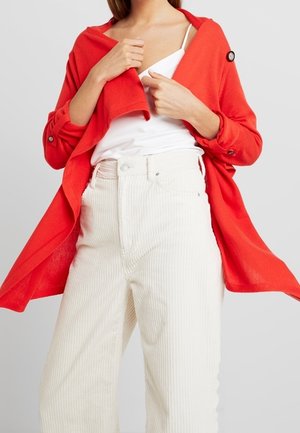 Femme portant un pantalon taille haute en velours côtelé blanc, un haut blanc et un manteau rouge vif fluide avec des détails à boutons.