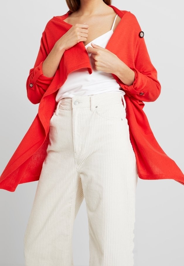 Femme portant un pantalon taille haute en velours côtelé blanc, un haut blanc et un manteau rouge vif fluide avec des détails à boutons.