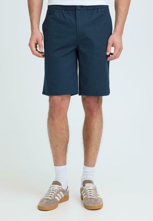 Mężczyzna w granatowych szortach do kolan, białych skarpetkach i beżowych trampkach w białe paski, stojący na jednolitym tle.