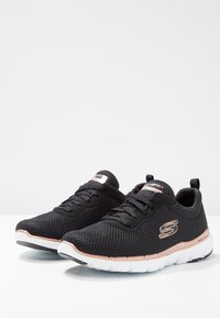 Svarta mesh-sneakers från Skechers med rosaguldiga detaljer, vita sulor, snören knutna, placerade sida vid sida på en ljus yta.
