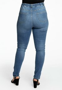 Jeans en denim bleu avec une taille élastique haute, coupe ajustée et deux poches arrière. Le tissu a une texture lisse et un léger élasticité.
