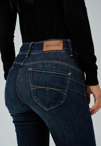 Jeans de mezclilla azul oscuro con una etiqueta de cuero que dice "SALSA JEANS", con bolsillos traseros estándar y costuras amarillas.